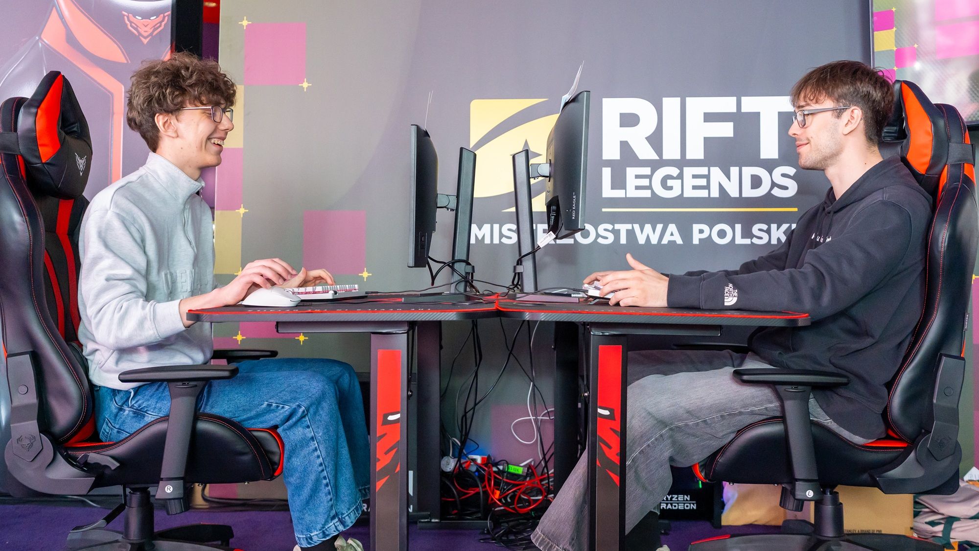 Dvaja hráči pri počítačoch pred stenou Rift Legends Majstrovstvá Poľska