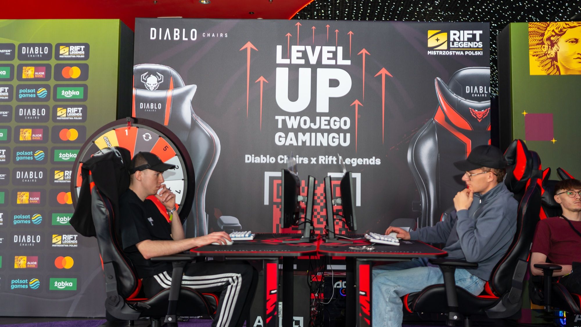 Dvaja účastníci pri stanovišti pred logami Rift Legends a Diablo Chairs